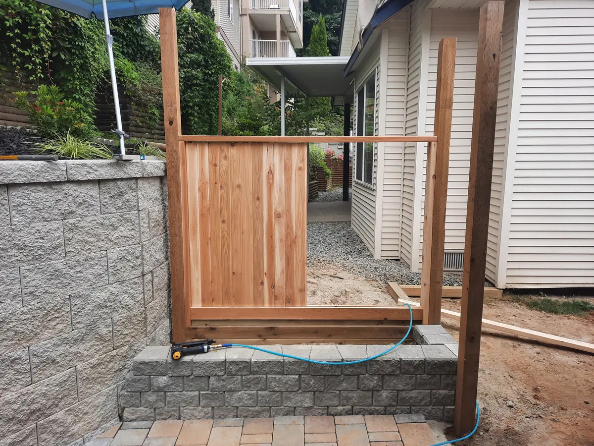 Cedar fence being built above a block planter, with boards going onto the frame between unstained posts, gravel path behind
