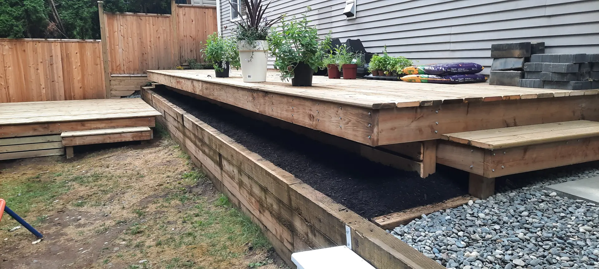 Finished wood deck with steps and a built-in planter bed edged with river rock, built by Blades Landscaping in Chilliwack