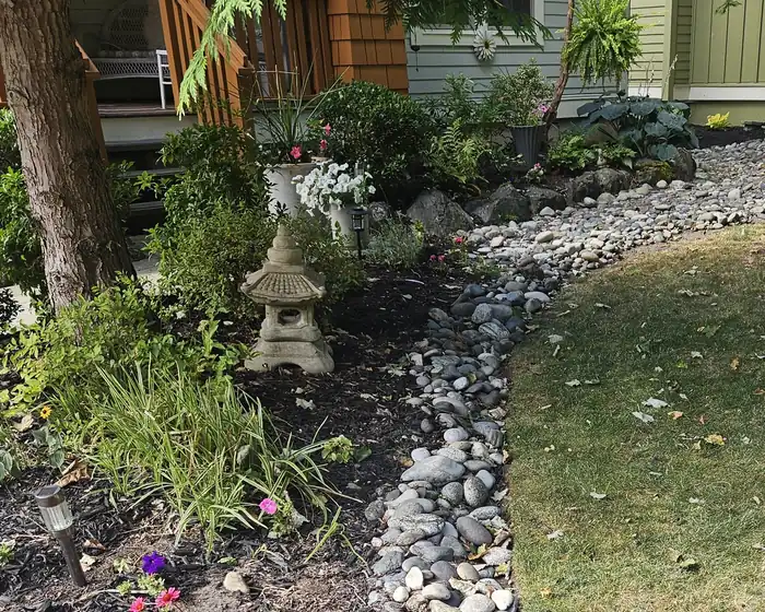 Before: front garden bed of dark mulch with flowers and a stone lantern, with a curving river rock border by a patchy lawn.