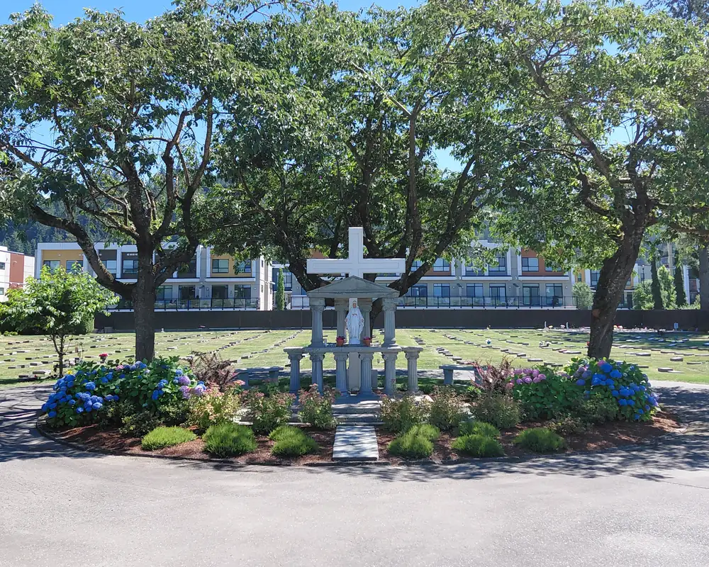 The same memorial garden with mulched beds of blue hydrangeas, flowering shrubs and grasses around the statue and cross.
