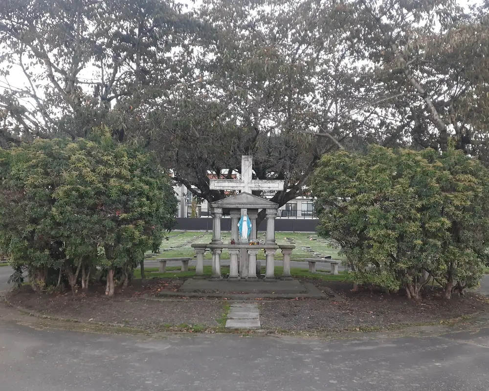 Memorial garden bed of bare soil with two large overgrown shrubs beside the statue and cross, ringed by a paved roundabout.