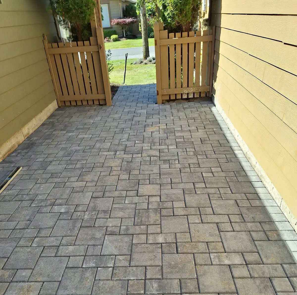 Newly laid grey paver patio extension in a narrow side yard, leading out to an open wood gate.