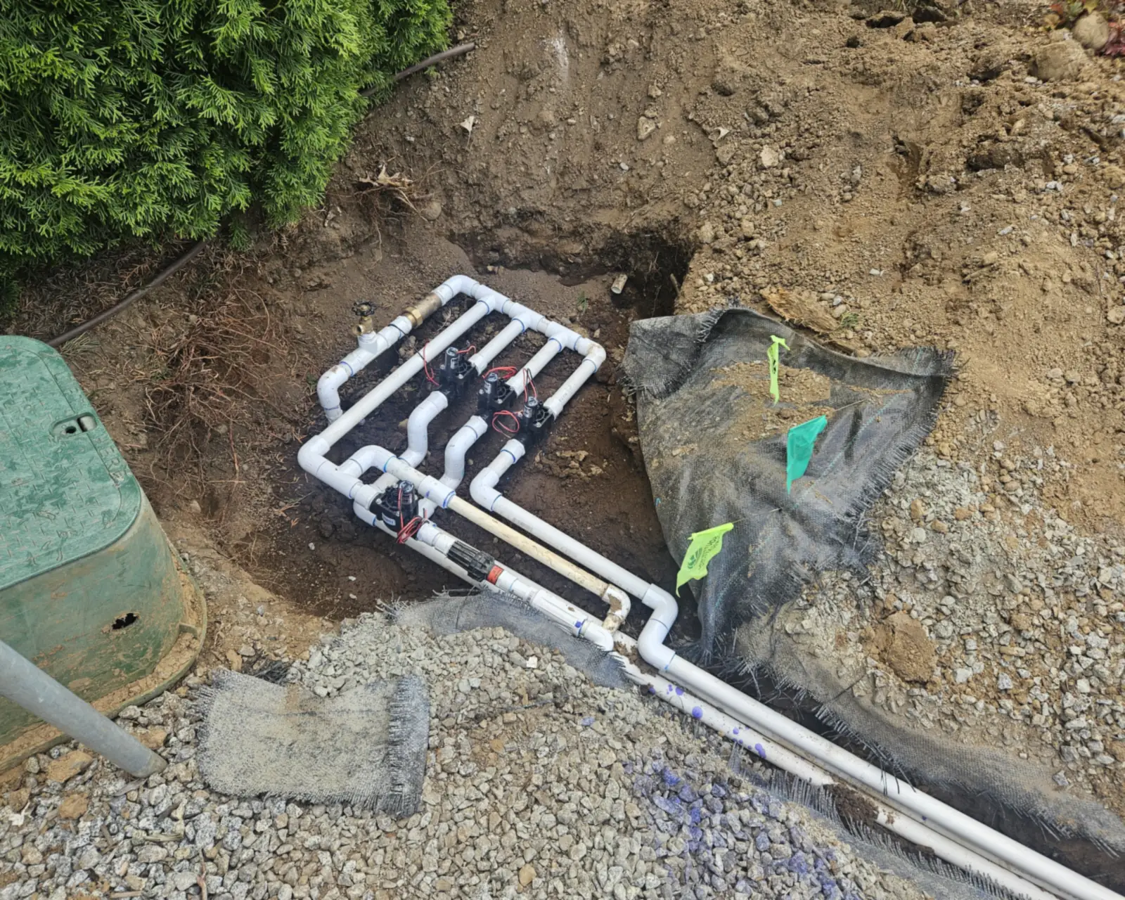 Sprinkler valve manifold with four zone valves and white PVC pipe, installed next to a green valve box