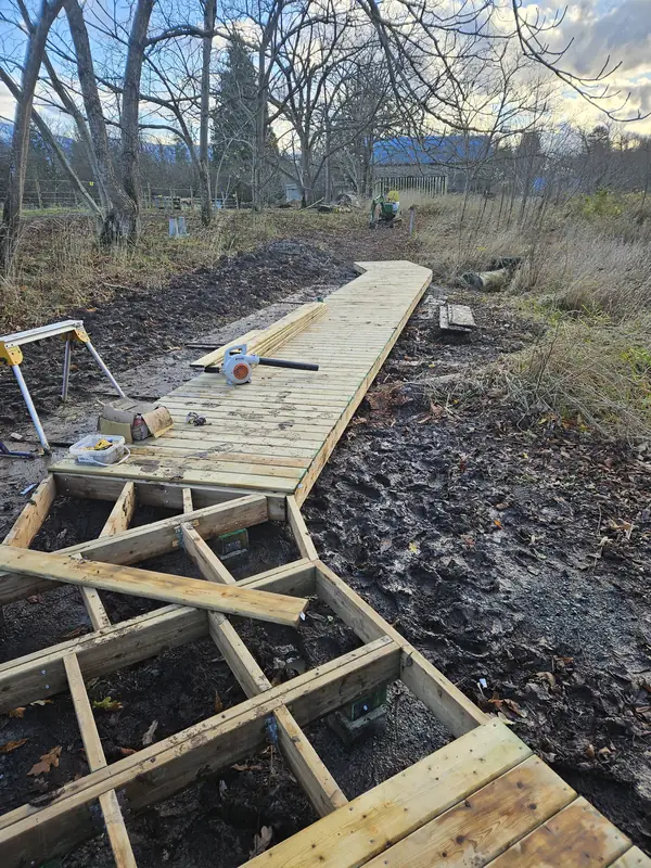 New boardwalk decking and open joist frame across wet, muddy ground in Agassiz, with tools on the deck and bare trees beyond